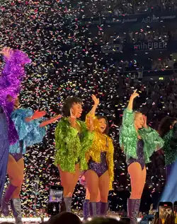 Vier Tänzerinnen stehen in bunten Kostümen auf der Bühne und winken im Konfetti-Regen ins Publikum.