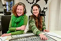 Zwei Frauen am Schaltpult eines Radios, beide tragen Kopfhörer, vor ihnen ein Radiomikrofon