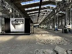 Zu sehen ist eine alte Industriehalle mit aufgerissenem Boden, hellem Licht aus Obrlichtern im Hallendach und eine provisorisch aufgebaute Bühne, an der ein Banner mit dem Logo von Museum Selma hängt.