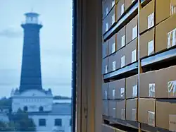 Zu sehen ist ein Archiv-Regal im klimatisierten Magazin von DOMiD. Die Fenster sind mit UV-Folie abgeklebt. Draußen ist der Helios Leuchtturm von Köln-Ehrenfeld zu sehen.