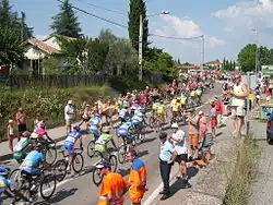 Seitliche Farbfotografie einer Landstraße mit vielen Radrennfahrern, die von links unten nach rechts oben radeln. Sie tragen verschiedene Trikotfarben und ein Nummernschild auf dem Rücken. An beiden Straßenseiten stehen jubelnde Menschen und rechts sind einige auf einer Mauer. Links oben ist ein Hang und zwischen den Bäumen steht ein Haus.