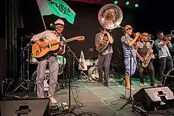 Eine Band tritt auf der Bühne auf mit Gitarre, Sousaphon, Trompete, Saxophon und Schlagzeug; ein Mitglied hält eine Fahne, und Mikrofone sowie Lautsprecher sind sichtbar.