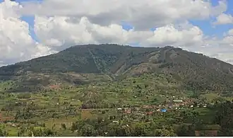 Ein niedriger, leicht bewaldeter Berg. Davor liegen locker verteilt kleine Landwirtschafts- und Weideflächen mit einzelnen Bäumen und Baumgruppen dazwischen. Rechts ist eine kleine Siedlung durch die eine Straße führt.