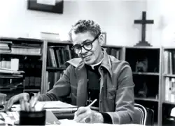 Eine ältere Pauli Murray sitzt mit dem Stift in der Hand an einem Schreibtisch. Im Hintergrund des Büros steht ein Kreuz auf dem Regal.
