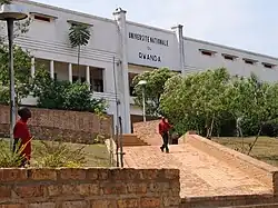 Blick auf einen über eine kurze Grünfläche führenden Weg mit einer Treppe am Ende vor dem Eingang des dreistöckigen Universitätsgebäudes. Über dem Eingang steht in Großbuchstaben Université nationale du Rwanda. Die unteren Stockwerke sind größtenteils durch Bäume verdeckt. Das erste Obergeschoss hat einen Laubengang und das Stockwerk darüber nur sehr kleine, schmale Fenster sowie ein Flachdach. Ein Mann kommt den Weg hinunter geschlendert.