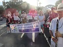 Mehrere Personen verschiedenen Alters stehen in einer Straße. Im Vordergrund halten zwei Frauen ein Banner mit der Aufschrift 45th Anniversary of the UpStairs Lounge Fire. Im Hintergrund halten zwei Männer Plakate mit den Aufschriften Unidentified White Male und Dr. Perry Lane Waters hoch. Am vorderen Rand des Umzugs steht ein Mann mit einer Klarinette, hinter ihm eine Frau mit einem Xylofon.