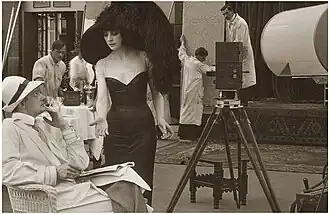 Urban Gad sitzt links in einem Stuhl mit Papieren, evtl. dem Drehbuch, auf dem Schoß. Asta Nielsen steht hinter ihm und hält fordernd die Hand nach vorne. Im Hintergrund arbeiteten mehrere Personen an den Kulissen.