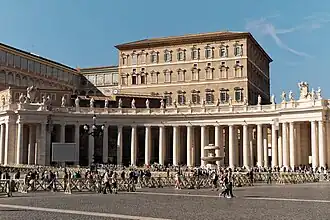 Farbfotografie von einem Platz mit Säulengang und Figuren darauf. Ein Springbrunnen ist auf der rechten Seite zu sehen und einige Menschen sind im Vordergrund. Hinter einer Absperrung am Säulengang hat sich eine lange Menschenschlange gesammelt. Über dem Säulengang ist links der obere Bereich eines Gebäudes mit vielen Fenstern zu sehen.