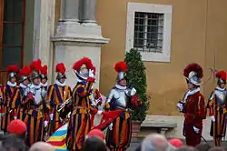 Frontale Farbfotografie einer Gruppe Gardisten mit Helmen, roten Federn und Halskrausen, die vor Zuschauern stehen. Drei von ihnen tragen ein Brustpanzer. Ein linker Gardist hält links eine Fahne und streckt mit der rechten Hand zwei Finger in die Höhe. Ein rechter Gardist steht mit einem Degen über seiner Schulter vor dem anderen und trägt eine dunkelrote Uniform.