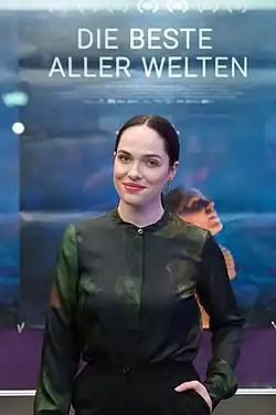 Verena Altenberger mit dunklen Haaren steht vor einem Filmplakat. Auf dem Plakat ist der Titel „DIE BESTE ALLER WELTEN“ in großen weißen Buchstaben oben zu lesen. Der Hintergrund des Plakats hat einen Farbverlauf von Blau zu Lila mit einigen verschwommenen Formen und Texten. Sie trägt ein dunkles, elegantes, langärmeliges Hemd mit einem dezenten Muster und schwarze Hosen. Das Bild wurde bei der Filmpremiere für den Film "Die Beste Aller Welten" in Wien 2017 aufgenommen.