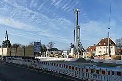 Man sieht Baugerät auf einer Baustelle hinter rot-weißen Absperrungen. Im Hintergrund sind Stadthäuser.