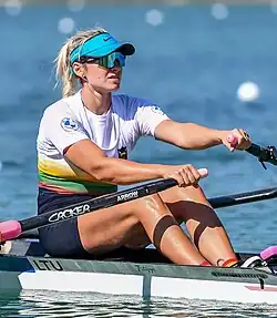 Viktorija Senkutė trägt das litauische Nationaltrikot über einem weißen T-Shirt und dunkelblaue Shorts. Das Boot ist weiß mit grauem Rand, die Ruder sind schwarz.