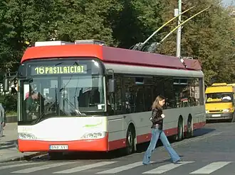 Frontale Farbfotografie eines rot-weißen Busses mit der Anzeige „16 Pasilaiciai“, der vor einem Zebrastreifen hält und Fahrgäste einsteigen lässt. Der Bus ist durch zwei Stangen mit der Oberleitung verbunden. Rechts läuft eine Frau über den Zebrastreifen. Im rechten Hintergrund steht ein gelber Kleintransporter.