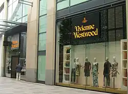 Seitliche Farbfotografie von einem Westwood-Geschäft in einem modernen Gebäude. Der gelbe Name Vivienne Westwood befindet sich mit gelbem Firmenlogo auf schwarzem Hintergrund und hängt über dem Eingang und im Schaufenster über sechs Schaufensterpuppen. Das gelbe Firmenlogo besteht aus einem Reichsapfel und dem ihn umkreisenden Planetenring.