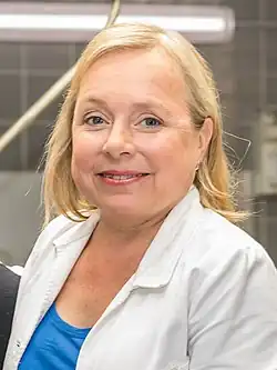 Christine Urspruch trägt einen weißen Laborkittel, darunter ein blaues Hemd. Die Umgebung deutet auf die Kulisse des Tatort Rechtsmedizin Labors hin, erkennbar an einer gefliesten Wand und mehreren Geräten, die unscharf im Hintergrund sichtbar sind. Die Szene wurde bei den WDR-Dreharbeiten zu Tatort Münster „Lakritz“-698 aufgenommen.