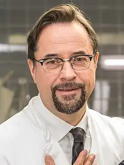 Jan Josef Liefers lächelt freundlich in die Kamera und trägt einen weißen Laborkittel, und eine gemusterte Krawatte. Er hat hellbraunes Haar und hält mit einer Hand den Laborkittel fest. Im Hintergrund ist eine Laborumgebung zu erkennen, mit gefliesten Wänden und einigen Geräten, die schemenhaft sichtbar sind. Das Foto entstand bei den WDR-Dreharbeiten zu Tatort Münster „Lakritz“ in der Rolle des Prof. Dr. Dr. Karl-Friedrich Boerne 2019.