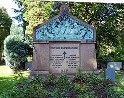 Von üppigem Grün umgebene breiter Grabstein aus rötlichem Granit in Form eines geschlossenen Tores. Ein giebelartiges Oberteil enthält ein Bronzerelief, das das Jüngste Gericht mit Christus als Weltenrichter auf einer Wolkenbank, umgeben von Heiligen und Engeln, darstellt.