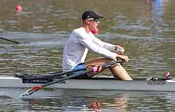 Clark Dean trägt eine schwarze Basecap, ein weißes, langärmliges Trikot und eine blaue Hose. Das Boot ist weiß, die Ruder sind schwarz.