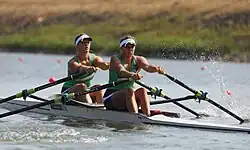 Die beiden Ruderinnen tragen eine Schirmmütze, grüne Trikots und schwarze Hosen.