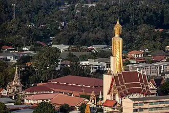 Die riesige goldfarbene Statue überragt alle Gebäude. Im Hintergrund ist bewaldetes Gebiet zu sehen.
