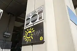 Detailaufnahme zweier rechteckiger Schilder, die an einer massiven Betonstütze eines gebäudes angebracht sind. Das obere Schild ist zweizeilig in schwarzen Lettern auf weißen Grund beschriftet: Obere Zeile New York City, untere Zeile als Logo die drei stilisierten Großbuchstaben DOT.