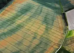 Luftbild mit den dunklen Bewuchsmerkmalen und Nachzeichnung des Erdwerkgrabens, darin Unterbrechungen durch frühere Erdbrücken