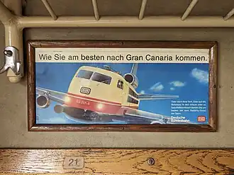 Ein Werbeplakat hängt in einem hölzernen Rahmen unterhalb einer metallenen Gepäckablage und oberhalb einer Holzleiste, auf der ein Aufkleber mit der Sitznummer 21 klebt. Das Plakat enthält die Überschrift „Die neue Bahn: Wie Sie am besten nach Gran Canaria kommen“. Darunter ist die Grafik einer Bundesbahn-Elektrolokomotive der Baureihe 103 zu sehen, die allerdings Flügel und Triebwerke eines Flugzeuges hat. Im Hintergrund blauer Himmel. Unten rechts auf dem Plakat steht kleiner: „Oder nach New York. Oder auf die Bahamas. In den Urlaub oder zu Geschäftsterminen starten Sie am besten mit dem Rail&Fly-Ticket der Bahn.“ Darunter etwas größer der Schriftzug „Deutsche Bundesbahn“ nebst dem zeitgenössischen Bundesbhan-Logo in rot und weiß.