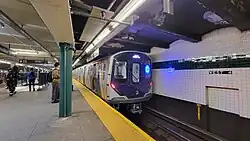 U-Bahn-Zug in unterirdischen Bahnhof von schräg vorne, Personen auf Bahnsteig, Hintergleiswand sichtbar, Zug zeigt blau leuchtendes LED-Liniensignal mit dem Großbuchstaben C, weiße LED-Schrift „168 ST / MANHATTAN“