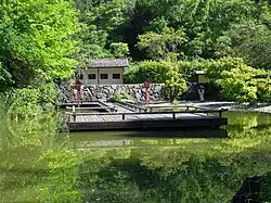 In einem kleinen See liegt einen Holzplattform, umgeben von Laternen, im Hintergrund eine Hecke und ein Wartehäuschen