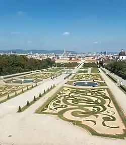 Farbfotografie in der Obersicht ornamentaler Beete mit fünf runden Wasserflächen. Auf dem mittleren Weg stehen Sträucher in Pyramidenform. Am linken und rechten Rand sind viele Bäume. Im Hintergrund sind eine Stadt und Berge.