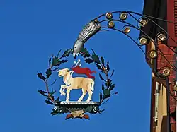 Ein am Gebäude befestigter Wirtshausausleger mit einem goldenen Lamm umrahmt von einem Eichenblätterkranz