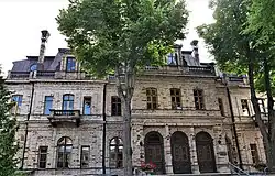 Frontale Farbfotografie eines zweigeschossigen Gebäudes mit hohen Rundbogeneingängen. Im Vordergrund eine Allee.