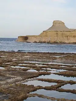 Im Hintergrund eine kegelförmige Felsformation, davor Meer, im Vordergrund rechteckige Salzpfannen