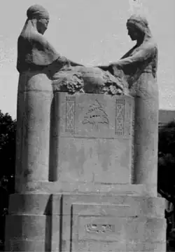 Darstellung aus Stein einer muslimischen Frau (links) und einer weiteren Frau (rechts) mit ausgestreckten Händen an einem altarartigen Tisch auf einem Sockel über einer Urne. Es sind geschnitzte auf den jeweiligen Truhen in Form eines kleinen Kreuzes und einer stilisierte Shahada zu erkennen von vorn in schwarzweiß.