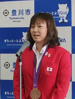 Yuka Katō wird nach der Rückkehr von den Olympischen Spielen in ihrer Heimatstadt empfangen und spricht in ein Mikrofon. Sie trägt eine rote Jacke über einer weißen Bluse und ihre Bronzemedaille ist an einem lila Band befestigt.