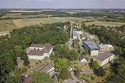 Luftbild, das im Vordergrund mehrere Gebäude, Wege und zahlreiche Laubbäume zeigt, und im Hintergrund Ackerflächen zeigt, die zum Teil durch Baumreihen getrennt sind.