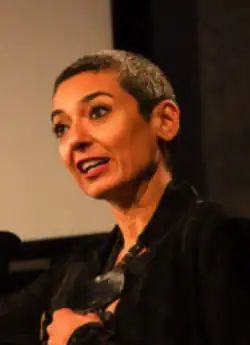 Zainab Salbi, kurze schwarz-grau-melierte Haare, trägt eine schwarze Jacke, sie steht und spricht stehend in ein Mikrophon