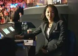 Das Bild zeigt Zhu Ying in einer halbnahen Einstellung. Sie sitzt an einem schwarzen Pult mit mehreren Displays und Unterlagen und hat sich im 90-Grad-Winkel zum Fotografen gedreht. Sie hat halblanges schwarzes Haar und trägt eine Halskette, ein weißes Shirt mit schwarzem Saum und ein schwarzes Jackett mit der Aufschrift „Strachan“ auf der linken Brust. Ihr rechter Arm ruht ausgestreckt auf dem Pult und das Licht fällt auf ihre rechte Kopfhälfte, während die linke Hälfte eher im Dunkeln liegt. Der Hintergrund ist weitgehend in dunklen Farben gezeigt, nur in der linken oberen Ecke gibt es einen bunteren Teil, in dem Teile des Snooker-Punlikums unscharf zu sehen sind.