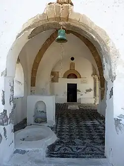 Das Foto zeigt einen Weißen Torbogen, durch den man unter einer Glocke in den Vorbau der Kirche auf die Türe zum Hauptraum blickt. Der Boden des Vorbaus besteht aus einem zweifarbigen Mosaik.