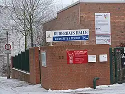 Das Ruderhaus ist mit Ziegelsteinen errichtet. Auf einer Tafel steht Bundesleistungszentrum und Landesleistungszentrum Rudern. Darunter ist die Tafel der Rudervereinigung mit den beiden gekreuzten Fahnen erkennbar. Das Gelände ist von einer Ziegelmauer umgeben, auf dem Boden liegt Schnee.