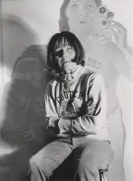 Maria Lassnig auf einem selbst fotografierten Schwarzweißbild von 1974, im Hintergrund das Gemälde Doppelselbstportrait mit Kamera
