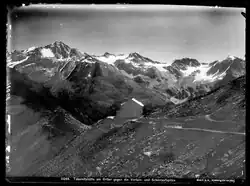 Schwarz-Weiß Fotografie einer Berglandschaft mit Felsen und Schnee. Etwa in der Bildmitte sieht man relativ klein ein Gebäude aus Stein mit reflektierendem Dach. Es steht an einem Abhang, ein Schotterpfad führt auf das Gebäude zu.