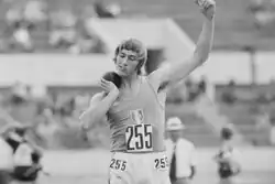 Das Schwarzweißfoto zeigt Le Roy beim Kugelstoßen in weißer Hose und grauem (in Farbe wäre es blau) Trikot. Vor seinem Versuch hält er die Kugel mit der rechten Hand gegen seinen Hals.