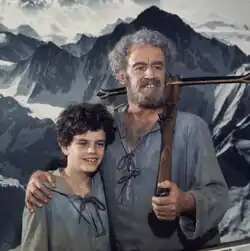 Zarli Carigiet mit Armbrust auf der Schulter in der Rolle des Wilhelm Tell, rechter Arm auf der Schulter seines Sohnes, beide in grauem Kapuzenhemd, blicken an der Kamera vorbei, Farbfotografie, quadratisch